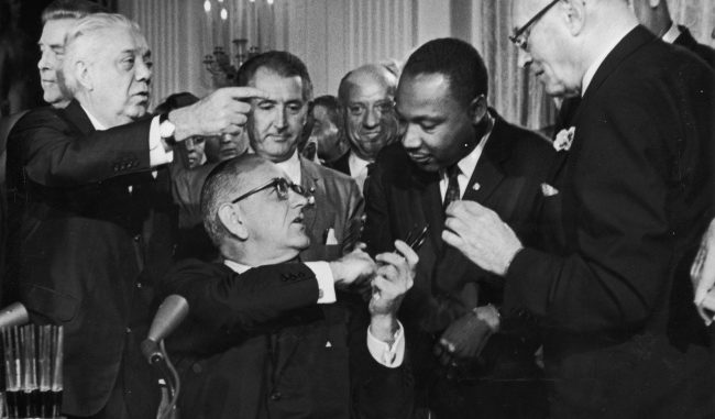 US President Lyndon B. Johnson shakes the hand of Dr. Martin Luther King Jr. (1929 - 1968) at the signing of the Civil Rights Act while officials look on, Washington DC. (Photo by Hulton Archive/Getty Images)
