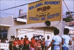 People gathered, some with drums, in front of wall with sign: New Jewel Movement (N.J.M.), National Secretariat, Let Those Who Labour Hold the Reins / credit: Center for Southwest Research, University Libraries, University of New Mexico