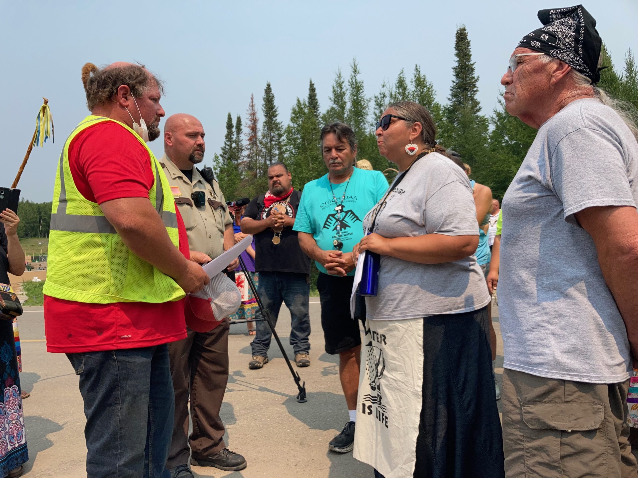 White Earth Tribal member and RISE Coalition co-founder Dawn Goodwin (second from right) serves an Enbridge representative with a cease-and-desist order for work on Line 3 under the Mississippi River. Clearwater County Sheriff Darin Halverson (second from left) flanks the representative while White Earth Tribal Council member Raymond Auginaush, Sr. (center) accompanies Dawn. Dawn and others established Camp Firelight near the headwaters of the Mississippi River at Coffee Pot Landing in Clearwater County, where Enbridge had set up pumping and drilling stations to bore under the river. Pipeline 3 or 5 is set to travel under the river, a cause for concern as it violates the sovereignty of Ojibwe people in the 1855 Treaty Territory XYZ and could pollute water for 20 million people who live downstream when the pipeline leaks. Pipeline leaks are common in the United States.