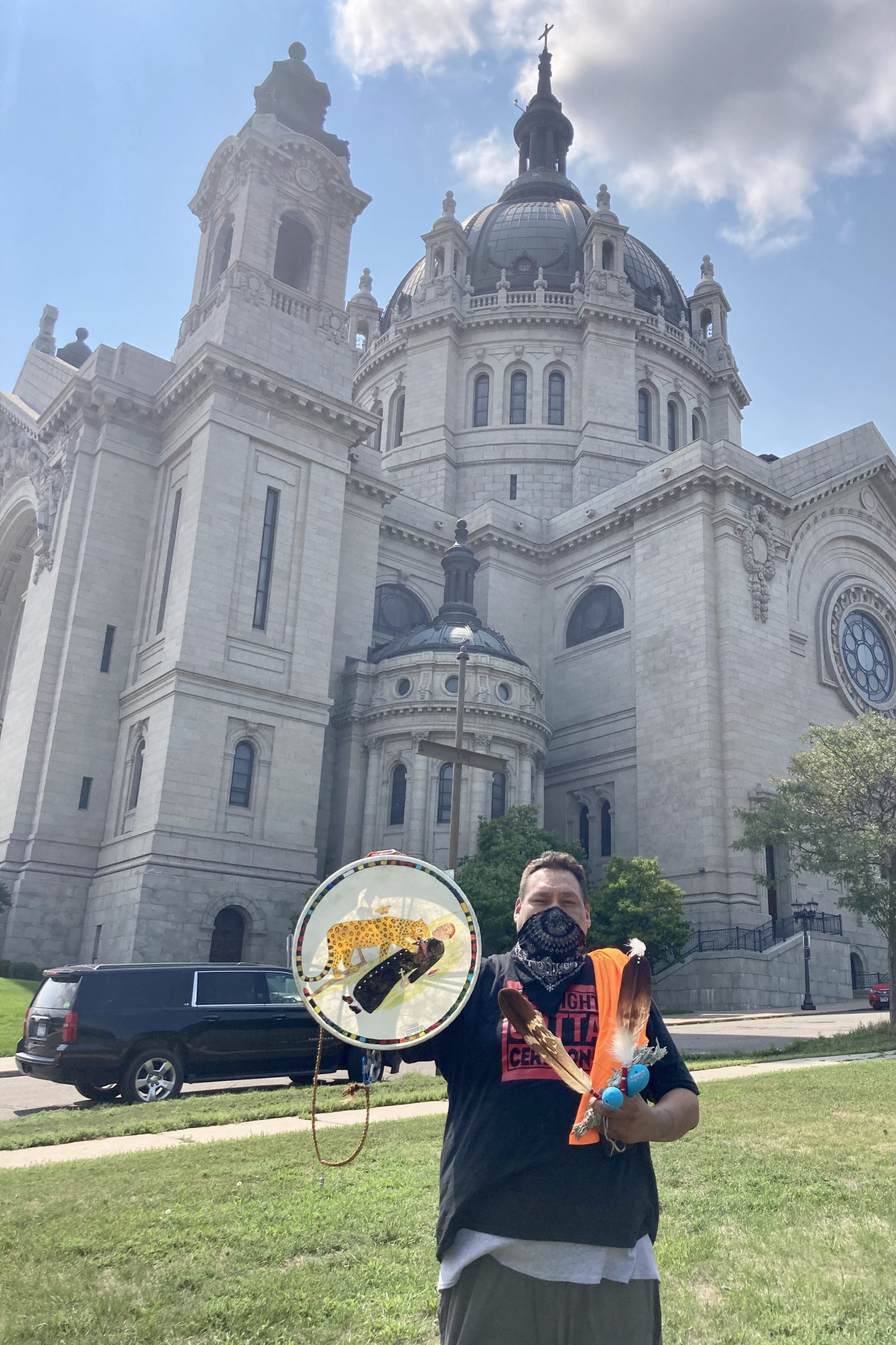 Hoka Wicasa holds feathers, sage and a hand drum depicting a jaguar eating the heart of a priest in front of a hearse and the Cathedral of Saint Paul at the end of the Treaty People Walk for the Water on August 25, 2021.