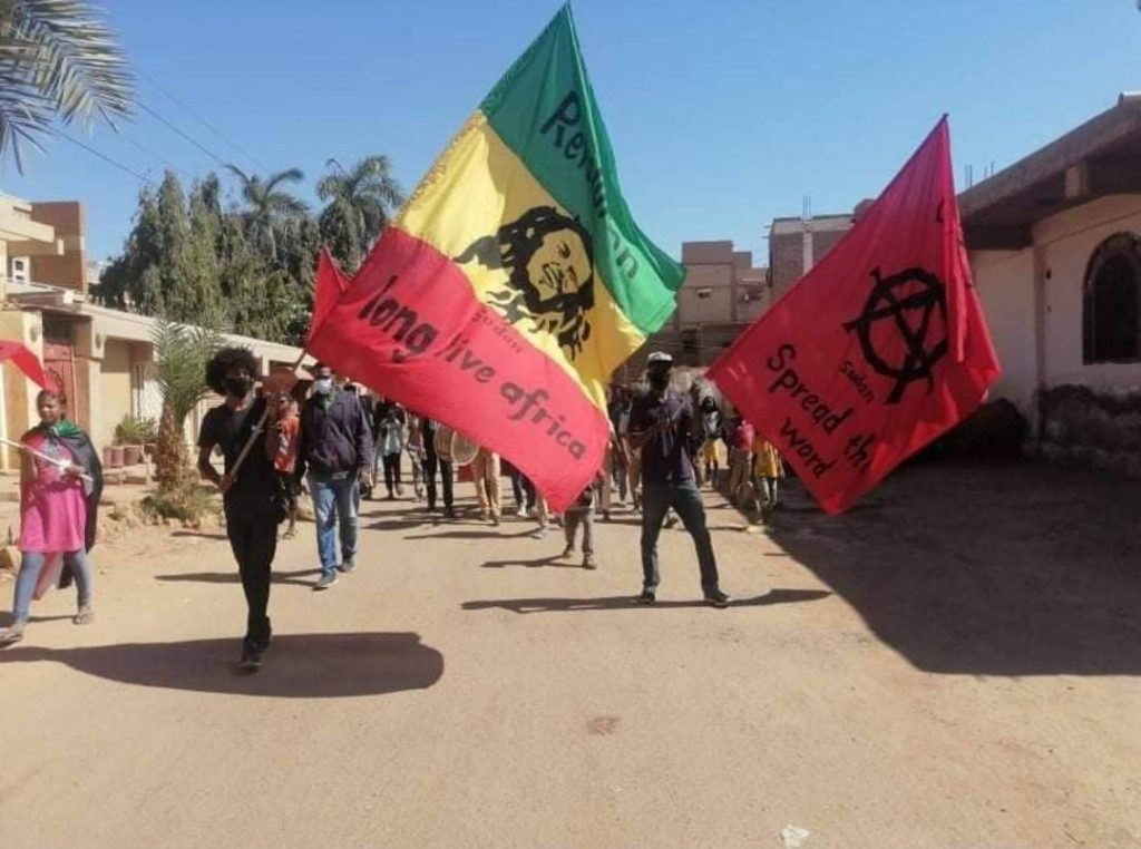 Protesters in Khartoum, Sudan, hold a red, yellow and green flag (center) that reads "Long live Africa." A red flag (right) contains the anarchist symbol and includes the phrase "Spread the word" / credit: Revolutionary masses of Sudan